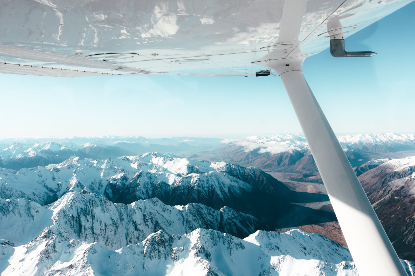 A wing-strut view from a light fixed-wing aircraft flying over snow-covered mountain ranges.