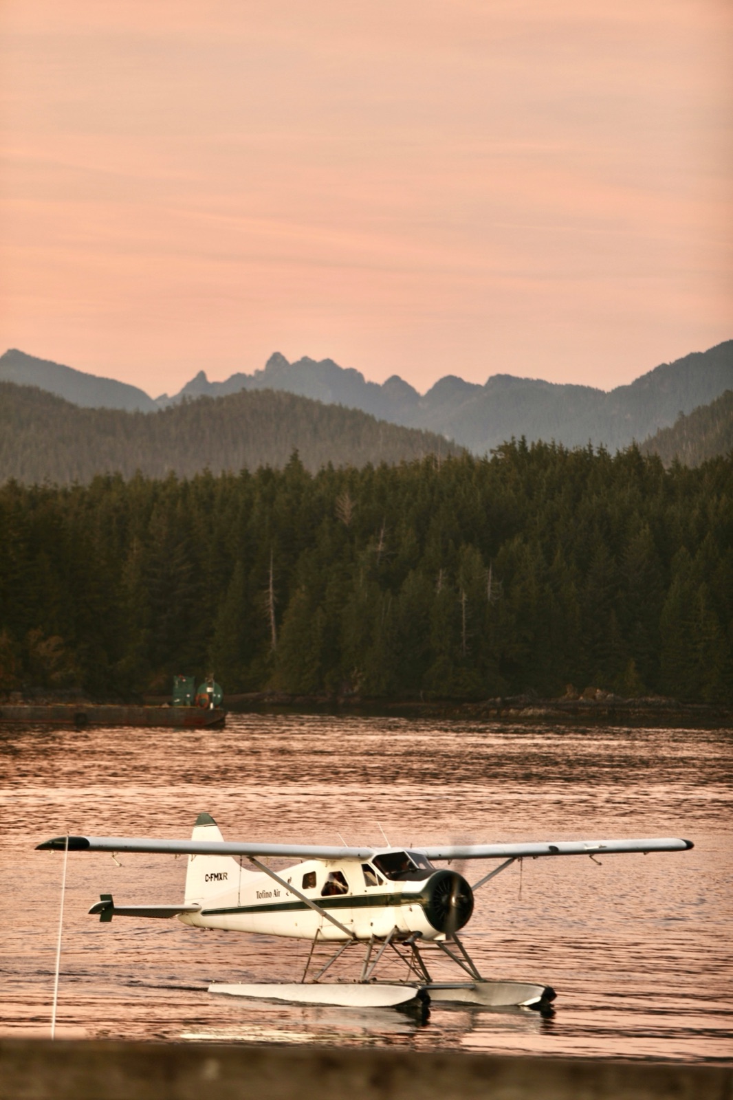 A DHC-2 Beaver floatplane on calm coastal water at dusk with forested mountains behind.